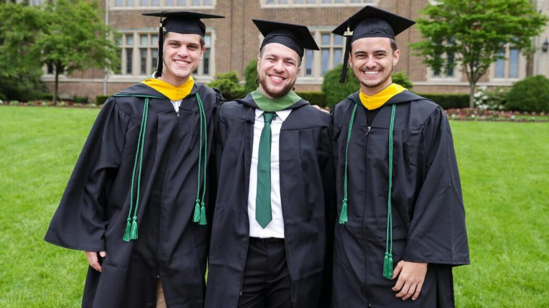 Three Mercyhurst graduates posing for a photo in graduation gowns in front of Old Main