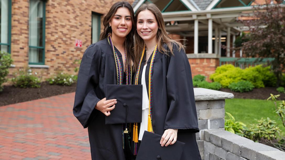 Two female Mercyhurst graduates, smiling for a photo outside Hammermill Library